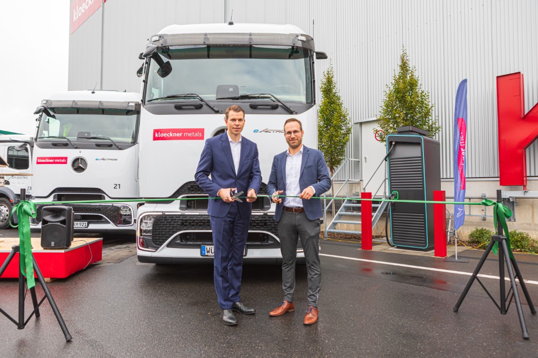 Zwei Männer in Anzügen schneiden vor einem großen weißen Lastwagen ein grünes Band durch und markieren damit ein Ereignis vor einem Gebäude an einem regnerischen Tag