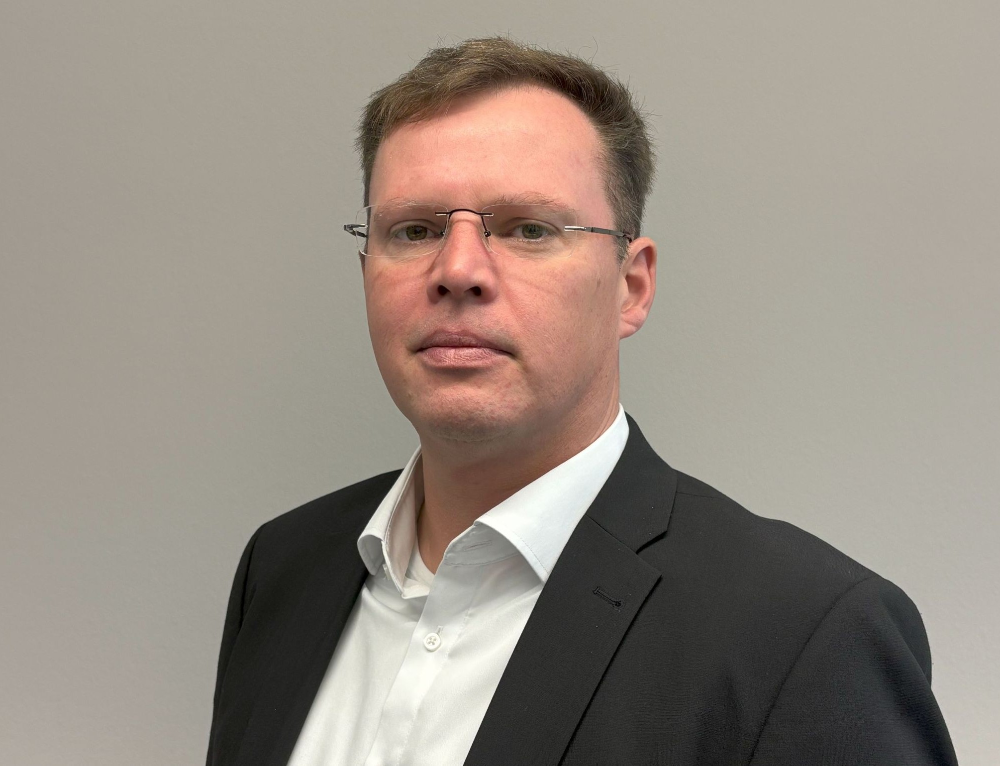 Ein Mann in schwarzem Anzug und weißem Hemd, der eine Brille trägt, steht vor einem schlichten grauen Hintergrund und blickt direkt in die Kamera.