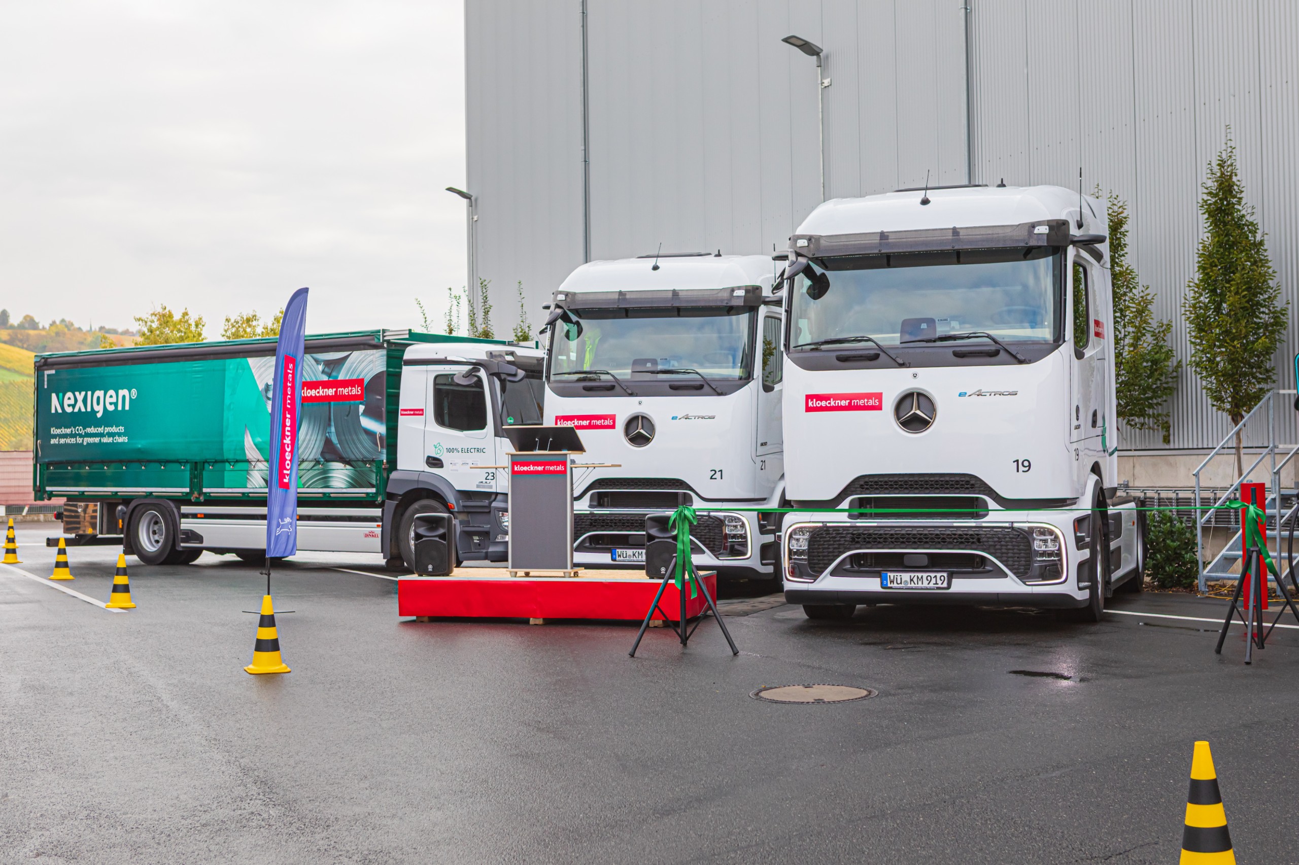 Drei weiße Lastwagen, die vor einem Gebäude geparkt sind, umgeben von Verkehrskegeln und einem grünen Lastwagen, mit Bannern und einem Podium in der Nähe.