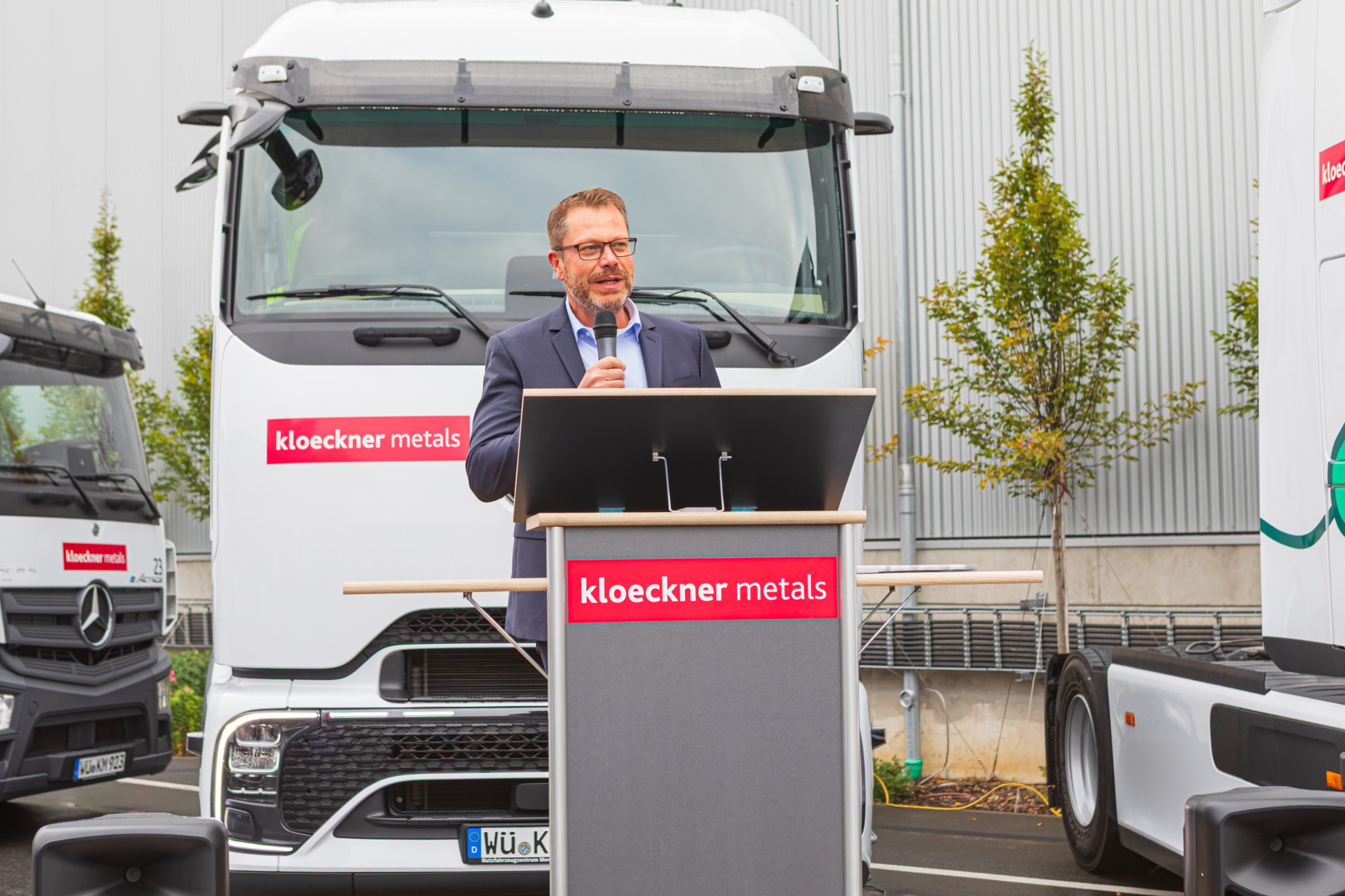Ein Mann spricht an einem Podium mit der Aufschrift „Kloeckner Metals“ vor Lastwagen mit dem gleichen Branding, die im Freien in der Nähe eines Gebäudes aufgestellt sind.