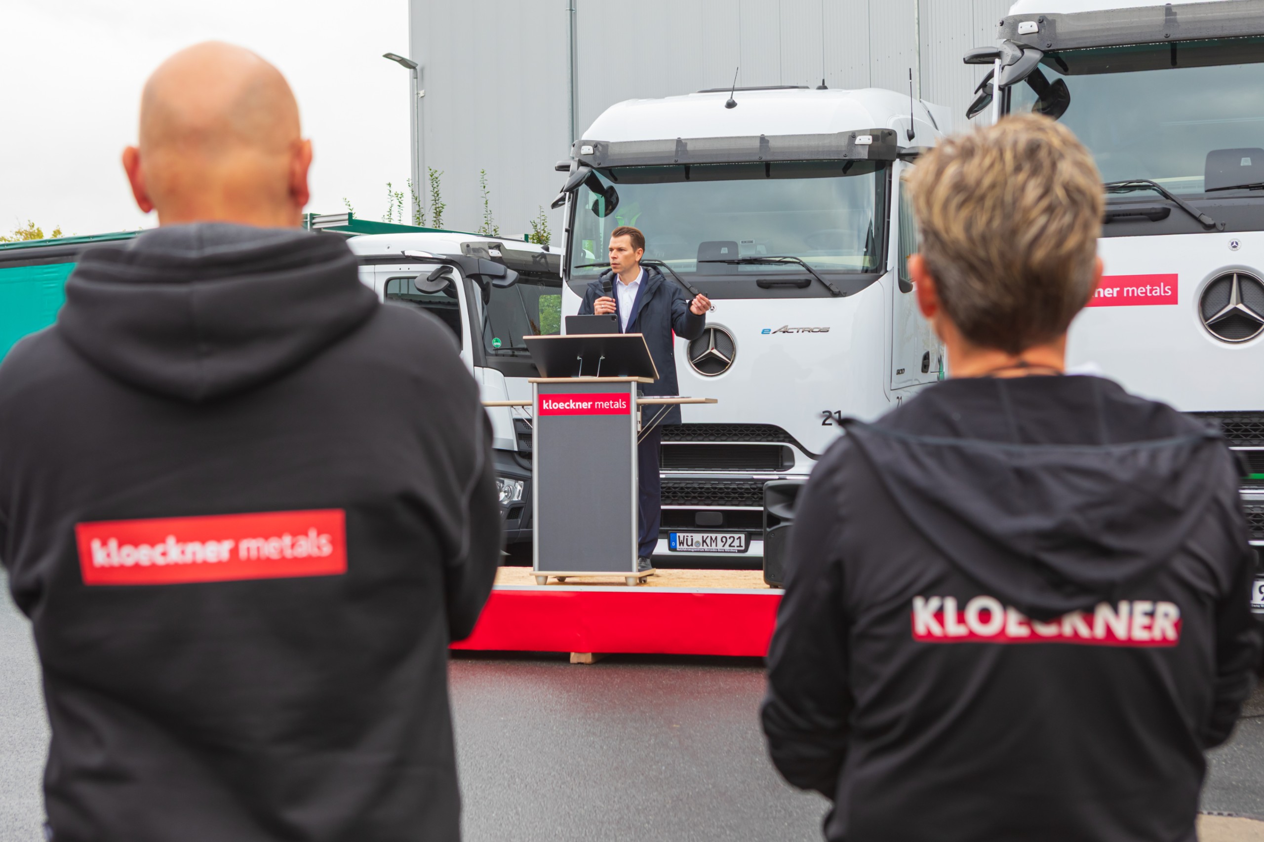 Eine Person spricht auf einem Podium vor Lastwagen, während zwei Personen in „Kloeckner Metals“-Kleidung im Vordergrund zuhören.