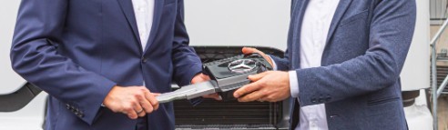 Zwei Personen in Anzügen halten einen großen Schlüssel vor einem Fahrzeug, im Hintergrund sind eine Treppe und ein Gebäude zu sehen.